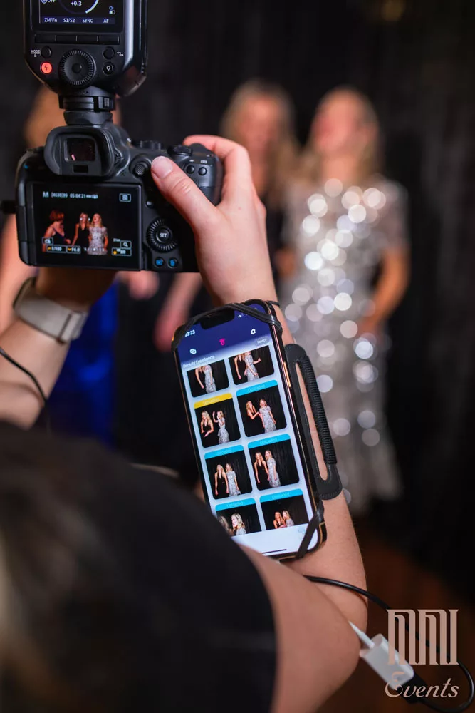 A photographer captures two people posing, with the camera screen and a connected smartphone displaying a corporate event photos in progress