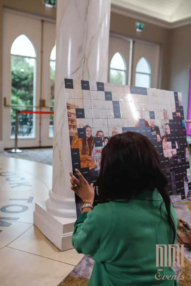 A woman in a green outfit is placing or adjusting a photo on a large mosaic display at a corporate event