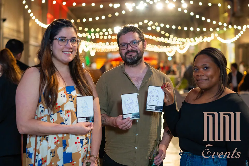 Three people are posing at a corporate event, each holding caricature portraits of themselves, with string lights creating a festive atmosphere in the background